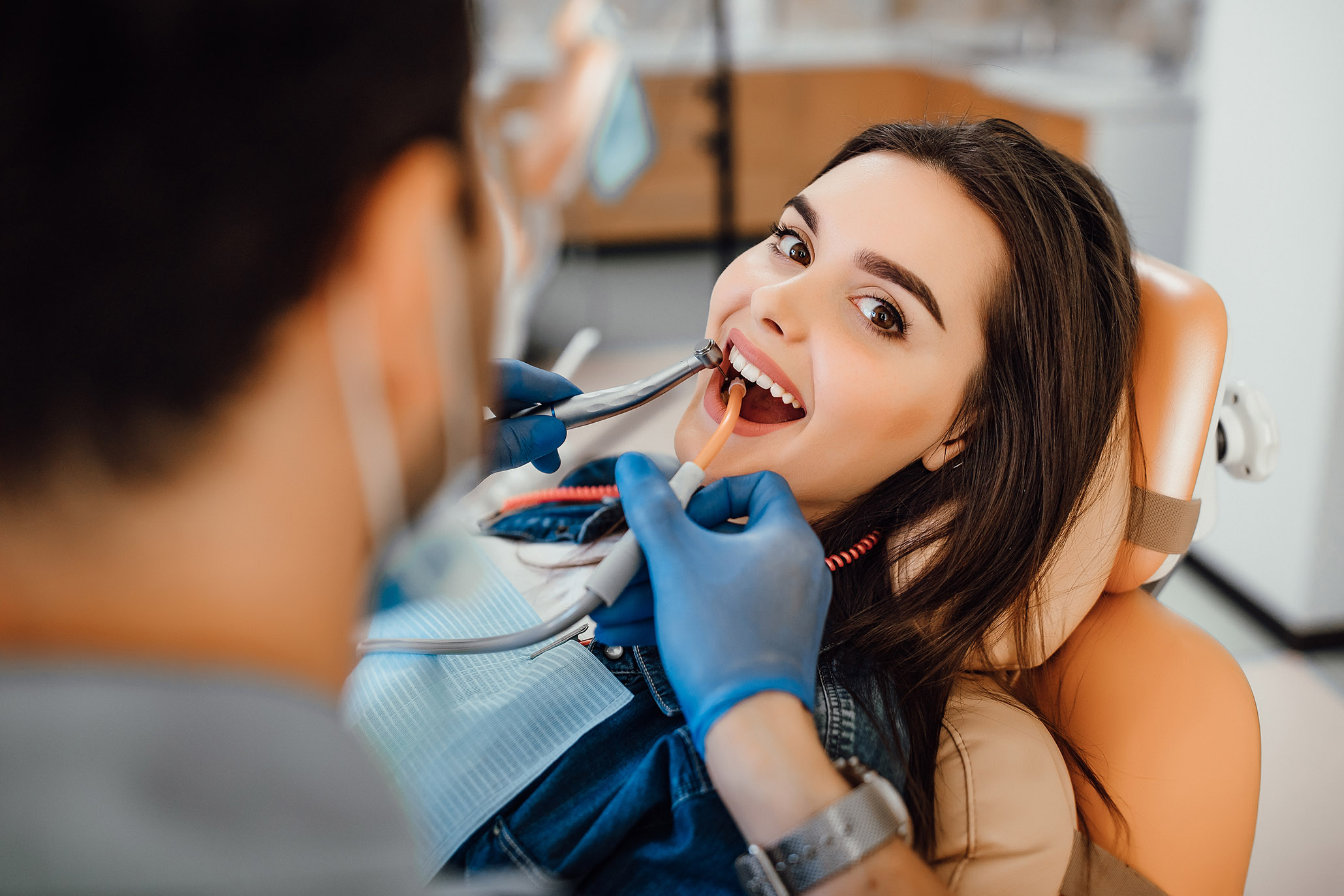 Young female patient visiting dentist office. Beautiful woman with healthy straight white teeth sitting at dental chair with open mouth during oral checkup while doctor working at teeth.